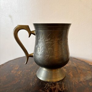 Peut inclure: Une chope en métal vintage avec une poignée décorative et des motifs floraux gravés. La chope a un aspect sombre et vieilli avec une poignée et une base de couleur laiton. La chope est présentée sur une table en bois sombre.