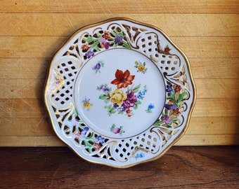 Peut inclure: Une assiette en porcelaine blanche avec un délicat motif ajouré orné de dorure. L'assiette présente un motif floral coloré avec des fleurs rouges, jaunes et violettes.