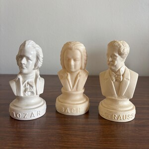May include: Three small busts of classical composers: Mozart, Bach, and Strauss. Each bust is a light beige color and features detailed facial features and clothing. The names of the composers are inscribed on the base of each bust.