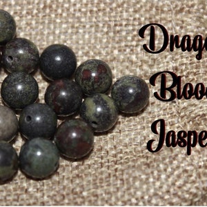 May include: A collection of round Dragon Blood Jasper beads in shades of green, brown, and black. The beads are arranged on a textured, tan surface. Text on the image reads "Dragon Blood Jasper".