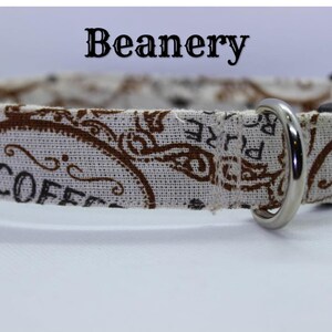 May include: A white and brown dog collar with a silver D-ring and a black plastic buckle. The collar has a vintage coffee design with the word "COFFEE" printed on it. The collar is labeled "Beanery".