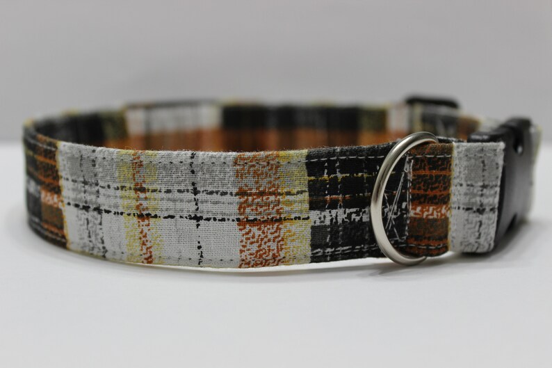 Op de afbeelding: Een bruin, zwart en wit geruit hondenhalsband met een zwarte gesp en een zilveren D-ring.