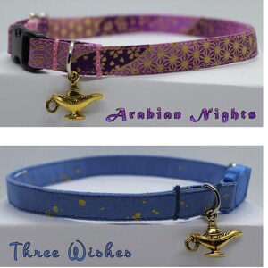 K&ouml;nnte beinhalten: Zwei Katzenhalsb&auml;nder mit goldenen Flaschengeist-Anh&auml;ngern. Das erste Halsband ist lila mit einem goldenen Blumenmuster und dem Text "Arabian Nights". Das zweite Halsband ist blau mit goldenen Punkten und dem Text "Three Wishes".