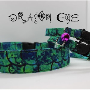 May include: Two green and blue cat collars with a black safety buckle and a bell. The collars have a mermaid scale pattern and the text "DRAGON EYE" is printed above the collars.