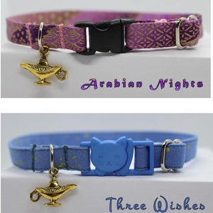 K&ouml;nnte beinhalten: Zwei Katzenhalsb&auml;nder mit einem goldenen Dschinnenlampen-Anh&auml;nger. Das erste Halsband ist lila mit goldenen Akzenten und dem Text "Arabian Nights". Das zweite Halsband ist blau mit goldenen Sternen und dem Text "Three Wishes".