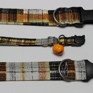 Op de afbeelding: Drie geruite kattenhalsbanden met zwarte gespen. De bovenste halsband heeft een zilveren ring. De middelste halsband heeft een zwarte kattenhanger en een oranje pompoenbel. De onderste halsband heeft een zilveren ring.