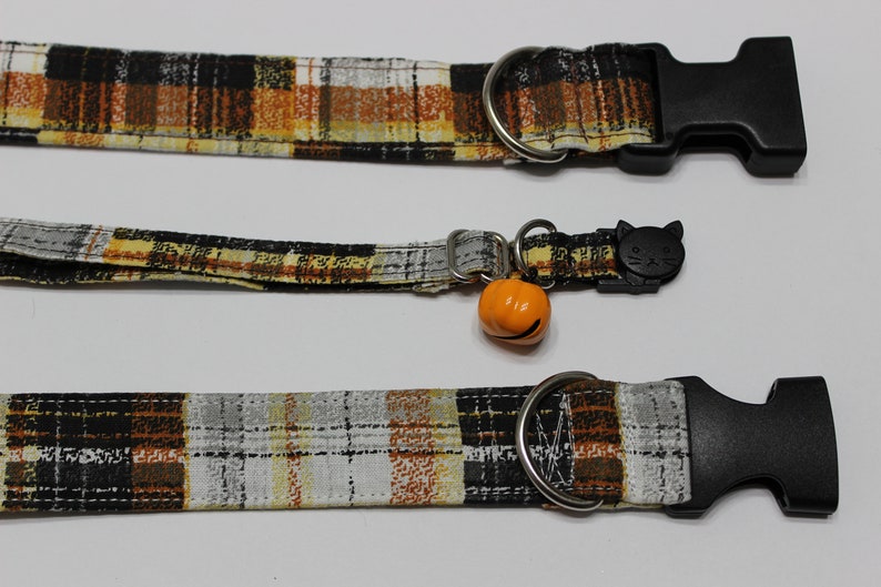 Op de afbeelding: Drie geruite kattenhalsbanden met zwarte gespen. De bovenste halsband heeft een zilveren ring. De middelste halsband heeft een zwarte kattenhanger en een oranje pompoenbel. De onderste halsband heeft een zilveren ring.
