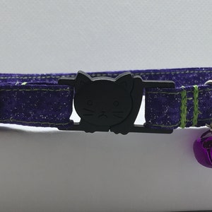 May include: Purple cat collar with a black cat face buckle and a purple bell. The collar has green stitching.