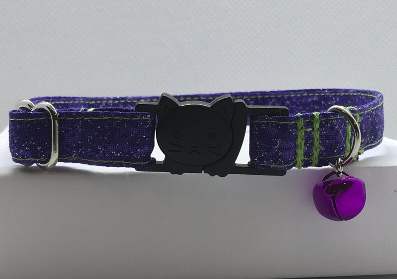 May include: Purple cat collar with a black cat face buckle and a purple bell. The collar has green stitching.