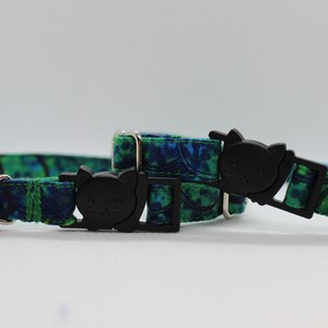 May include: Two green and blue patterned cat collars with black cat-shaped buckles and a bell on each. One bell is pink and the other is green.