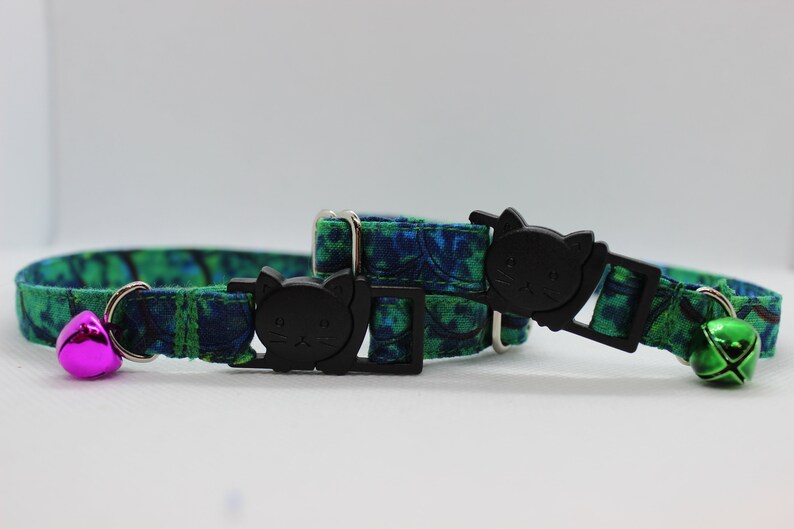 May include: Two green and blue patterned cat collars with black cat-shaped buckles and a bell on each. One bell is pink and the other is green.