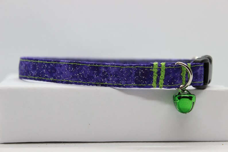 May include: Purple cat collar with green trim and a green bell. The collar has a black buckle.