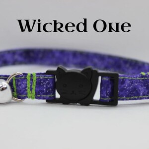 May include: Purple and green cat collar with a black cat charm and a silver bell. The collar has a white snowflake pattern. The text "Wicked One" is written above the collar.