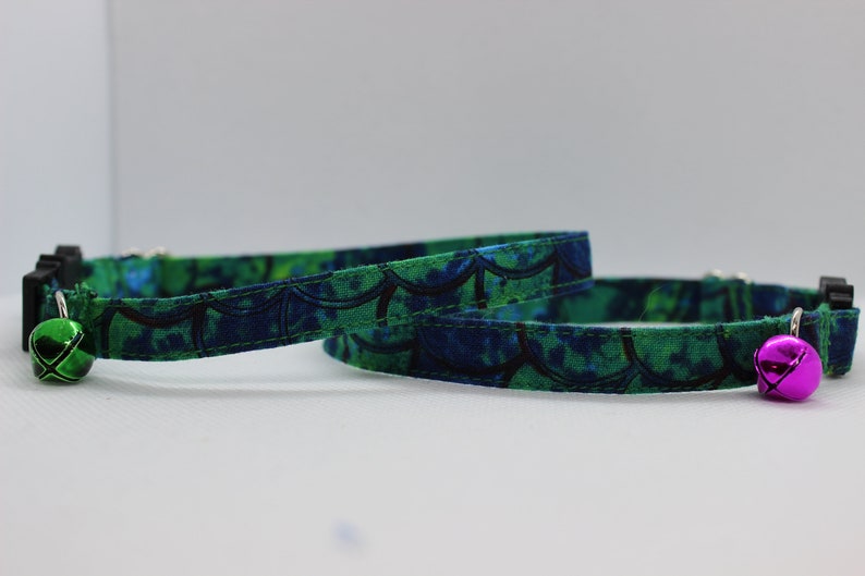 May include: Two green and blue patterned cat collars with a bell on each end. One bell is green and the other is pink.