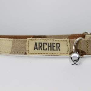 May include: Camouflage cat collar with a beige plastic buckle and a silver bell. The collar has the name "ARCHER" printed on a beige label. The pattern is a mix of tan, brown, and beige.