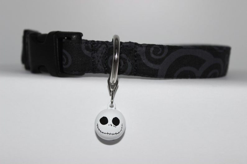 May include: Black and gray patterned dog collar with a silver D-ring and a white bell with a black Jack Skellington face.