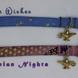 K&ouml;nnte beinhalten: Zwei Katzenhalsb&auml;nder mit einem goldenen Dschinnenlampen-Anh&auml;nger. Das obere Halsband ist blau mit goldenen Glitzer und tr&auml;gt die Aufschrift "Three Wishes". Das untere Halsband ist lila mit goldenen Glitzer und tr&auml;gt die Aufschrift "Arabian Nights".