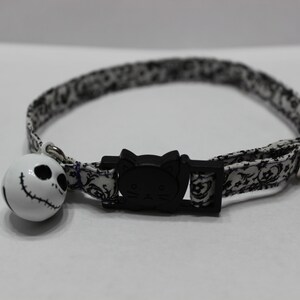 May include: A white and black patterned cat collar with a black cat face buckle and a white bell with a black Jack Skellington face.