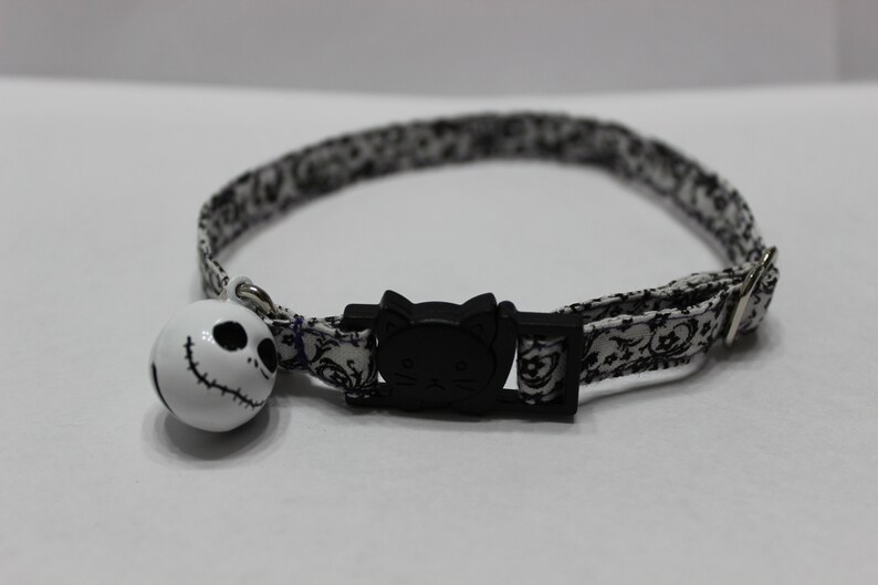 May include: A white and black patterned cat collar with a black cat face buckle and a white bell with a black Jack Skellington face.