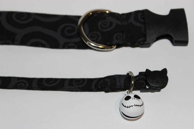 May include: Black cat collar with a black and gray swirl pattern and a silver buckle. The collar has a black cat charm and a white bell with a Jack Skellington face.
