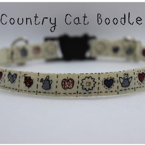 May include: A cream-colored cat collar with a black plastic buckle. The collar is decorated with a repeating pattern of red hearts, blue angels, and other country-themed designs.