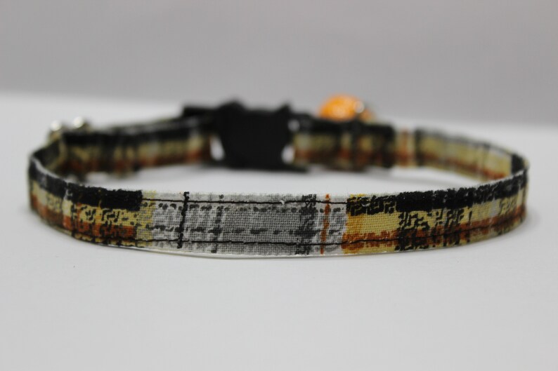 Op de afbeelding: Een zwarte, oranje en witte geruite kattenhalsband met een veiligheidssluiting en een bel.
