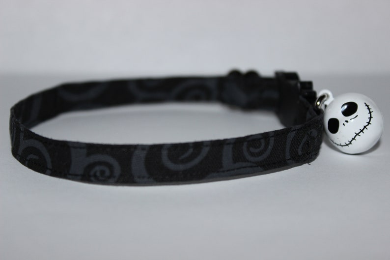 May include: Black cat collar with a white Jack Skellington bell charm. The collar has a black and gray swirl pattern.