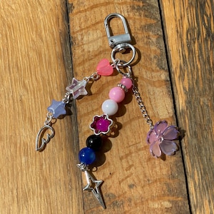 May include: A silver-toned metal keychain featuring three dangling strands of beads and charms. The strands include pink, white, purple, black, and blue beads, along with star-shaped charms, a heart, a wing, a flower-shaped bead, and a metal star.