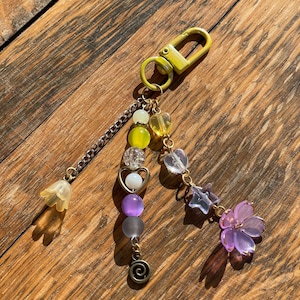 May include: A decorative keychain with a yellow clasp and a chain. The chain is adorned with a variety of beads in yellow, purple, and clear colors, along with a flower and star charm. The keychain is set against a wooden background.