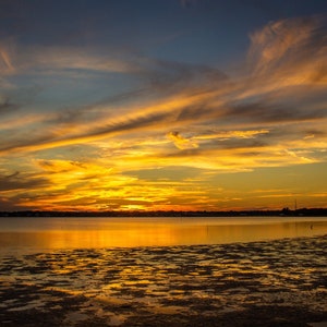 May include: A scenic sunset over a body of water with a golden sky and clouds reflecting in the water. The water is calm and still, with a sandy shoreline in the foreground.