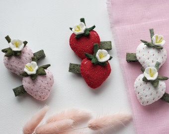 Puede incluir: Tres pares de pinzas para el pelo de fresa de fieltro. Cada par presenta dos fresas con una flor blanca en la parte superior. Las fresas son rojas, rosas y blancas.