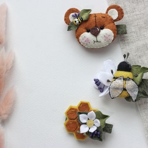 May include: Three felt hair clips featuring a bee with a white flower, a honeycomb with a white flower, and a brown bear with a bee on its head.