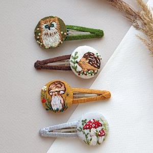 May include: Four hair clips with embroidered designs. The clips are brown, green, yellow, and gray. The designs are an owl, a hedgehog, a mushroom, and a mushroom with red dots.