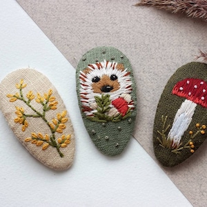 Może przedstawiać: Trzy haftowane spinki do włosów z tkaniny z motywami natury. Jedna spinka przedstawia żółty kwiat, druga jeża, a ostatnia czerwony i biały grzyb.