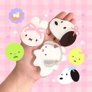 May include: A collection of five colourful cartoon character acrylic charms. The charms feature a white bunny with pink ears and a pink bow, a white dog with black ears and a pink bow, a white bunny with grey ears and a grey headband, a green cartoon face with a frown, and a yellow cartoon face with a frown.