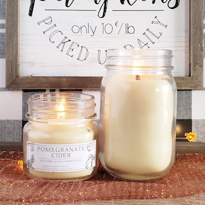 May include: Two lit glass jar candles with a wooden sign in the background. The sign reads "Farm Fresh Pumpkins Picked Daily" and "only 10¢/lb". The smaller candle has a label that says "Pomegranate Cider" and "Autumn at the Farmhouse".