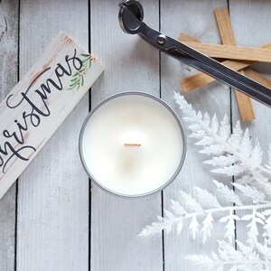 Peut inclure: Une bougie blanche dans une boîte en étain argentée avec une mèche en bois. La bougie est posée sur une surface en bois blanc avec une pancarte qui dit "Joyeux Noël" et une paire de coupe-mèches de bougie.