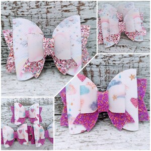May include: Two white and pink hair bows with a white and pink glitter overlay. The bows have a white background with a pink and blue watercolor design. The bows are layered with a white sheer fabric.