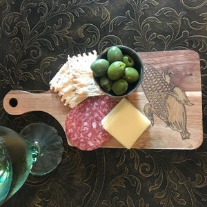 May include: A wooden charcuterie board with a corn design, holding green olives, crackers, salami, and cheese. A wine glass is in the foreground. The items are arranged on a dark, patterned surface.