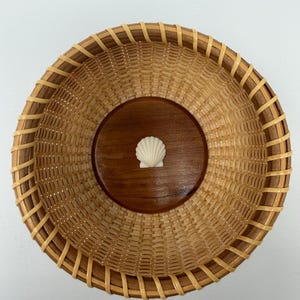 May include: A round woven basket featuring a smooth, dark brown wooden base with a small, white, fan-shaped seashell centred on the bottom. The sides are constructed from light tan woven reeds with a reinforced, darker brown wooden rim.