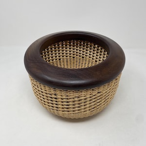May include: A handcrafted basket with a woven, light brown body and a dark brown wooden rim. The basket's design features a circular shape, perfect for storage or display. The rim is approximately 10 inches in diameter.