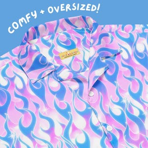 May include: A button-down shirt with a vibrant flame pattern in shades of pink, blue, and white. The shirt features a collar and buttons down the front. The text "COMFY + OVERSIZED!" is written at the top. A label reads "SILLY GOOSE" and "WORKSHOP & GOODS".