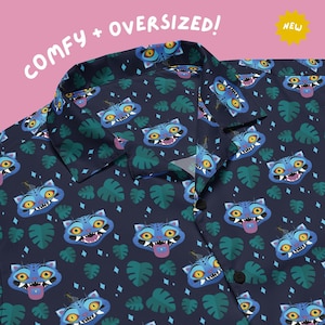 May include: Navy blue button-down shirt with a repeating pattern of blue cat faces and green monstera leaves. The shirt has the words "COMFY + OVERSIZED!" and a "NEW" sticker in the top corners.