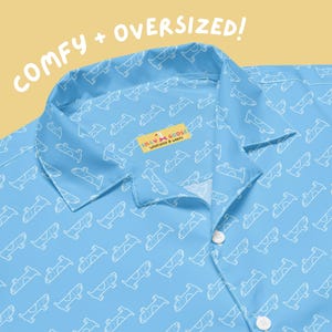 May include: Light blue button-up shirt with a white goose pattern. The shirt has a collar and two white buttons. The text "COMFY + OVERSIZED!" is in the upper left corner. The label reads "SILLY GOOSE WORKSHOP & WARES".
