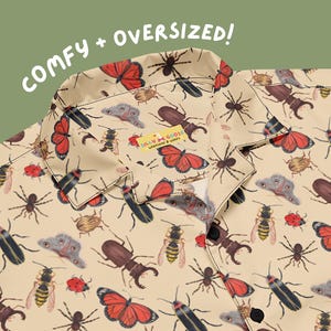 May include: A beige button-down shirt featuring a repeating pattern of various insects including butterflies, spiders, beetles, and wasps. The collar area displays a small yellow tag with the text Silly Goose Workshop & Wares.