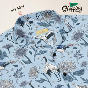 May include: A light blue button-down shirt featuring a repeating pattern of white and black cranes and dark blue chrysanthemum flowers. The shirt has a notched collar, black buttons, and a yellow inner neck label that reads silly goose.