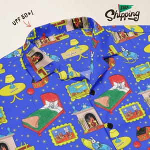 May include: A bright blue button-down shirt featuring a repeating pattern of rabbits in beds, fireplaces, lamps, and framed pictures. The collar tag reads "silly goose" and the fabric is labeled with "UPF 50+!" text printed on the upper left side.