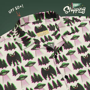 May include: A short-sleeved button-down shirt with a cream background featuring a repeating pattern of black moth-like creatures with red eyes, dark green pine trees, and grey flying saucers with purple light beams. The shirt has a collar.