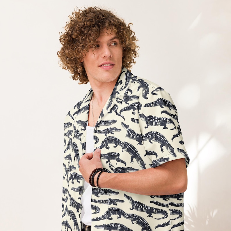 May include: A short-sleeved button-down shirt in a light cream color featuring an all-over repeating pattern of dark navy blue alligators. The shirt has a classic collar and is worn over a plain white undershirt, showcasing a casual style.