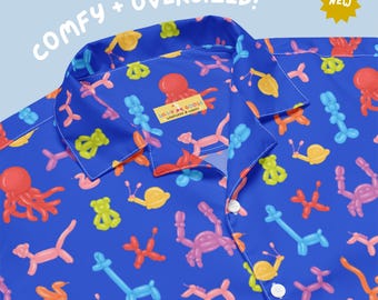 May include: A blue button-down shirt featuring a repeating pattern of colorful balloon animals including octopuses, snails, and dogs. The shirt has a collar and white buttons. Text on the image reads COMFY + OVERSIZED! and NEW. A label reads SILLY GOOSE.